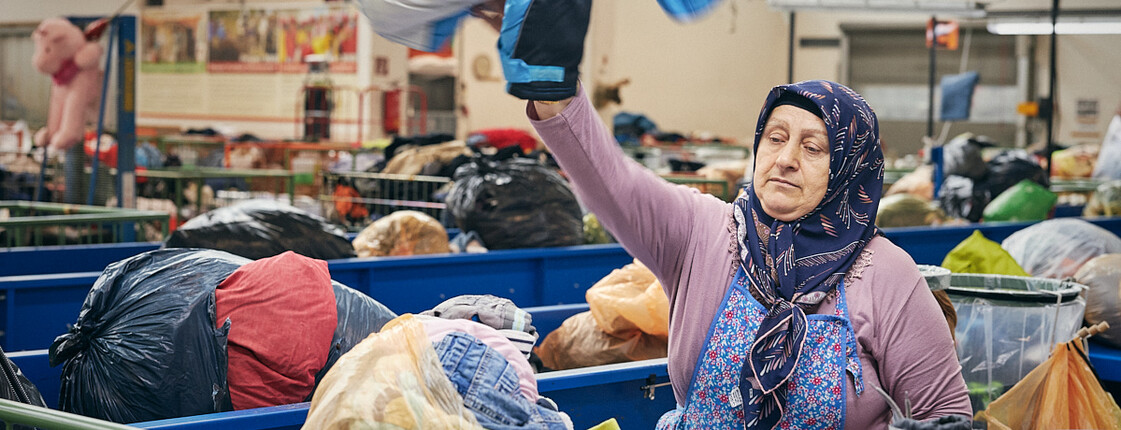 Eine ältere carla Vorarlberg Mitarbeiterin mit Kopftuch, die Gebrauchtkleider im Kleidersortierwerk sortiert und für den Wiederverkauf in den carla Secondhand Shops aufbereitet