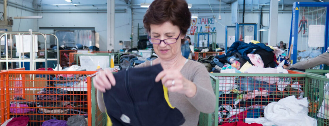 Eine carla Mitarbeiterin, die kurze braune Haare hat und eine graue Bluse trägt, sortert Kleidung. Sie ist etwa mittleren Alters.