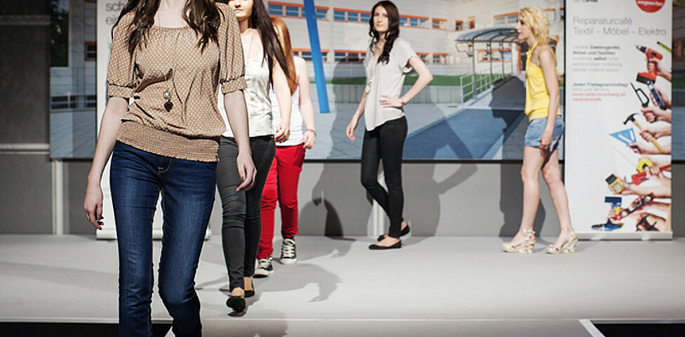 carla Modeschau: Fünf weibliche Models laufen elegant auf dem Laufsteg und präsentieren sich und die von ihnen getragene Mode. Sie tagen unter anderem beige und weiße Oberteile. Bis auf eine Dame tragen alle lange Hosen. 