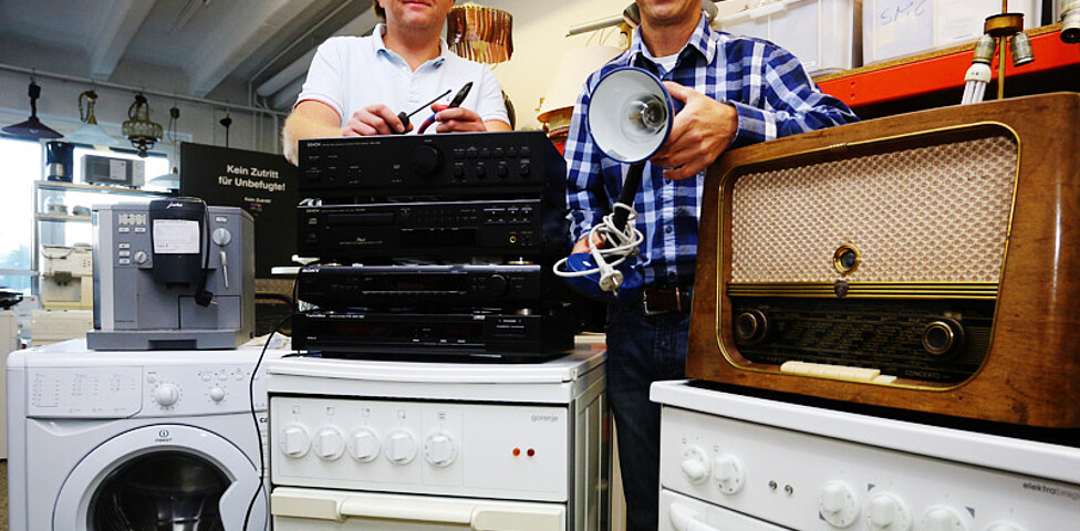 Zwei Männer mittleren Alters stehen hinter defekten Elektogeräten. Das sind unter anderem Lautsprechanlagen und weißen Waschmaschienen. 
