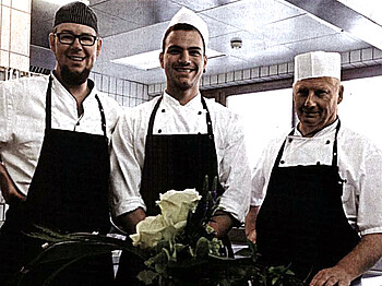 Drei Köche unterschiedlichen Alters sehen in die Kamera. Sie tragen weiße Arbeitskleidung mit schwarzen Schürtzen und stehen in der Küche. 
