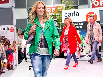carla Modeschau: Models unterschiedlichen Alters laufen schick gestylt über den weißen Laufsteg. Dabei tragen die Teilnehmerinnen unterschiedliche Farben wie grün, rot und beige. 