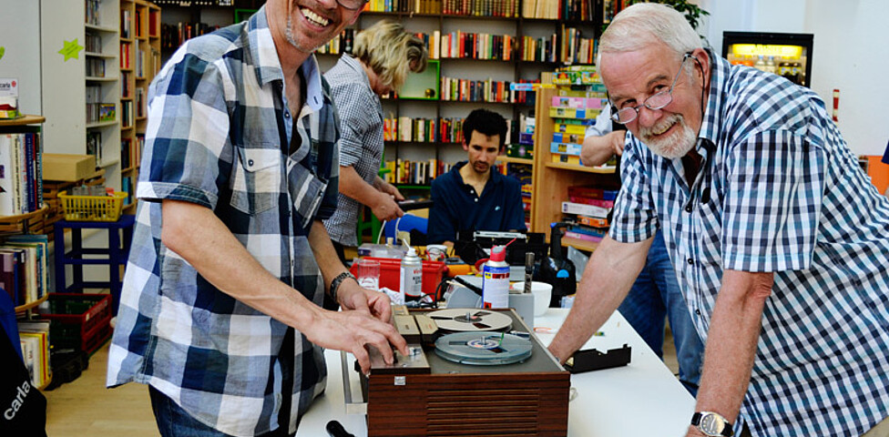 Ein älterer Herr und Mann mittleren Alters, die beide karierte Hemden tragen, reparieren ein altes Aufnahmegerät, das auf einem weißen Tisch steht. Die Stimmung ist entspannt und beide lachen. 