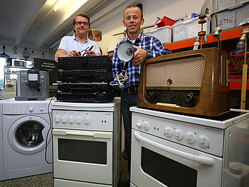 Zwei Männer mittleren Alters reparieren defekte Elektrogeräte wie Waschmaschinen, Herde und Lautsprecheranlagen. 