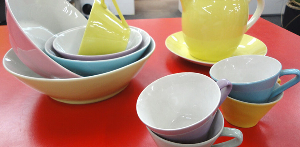 Farbiges Geschirr steht auf einem roten Tisch. Es sind Tassen, Kürge und Schalen. 