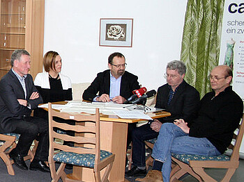 Fünf Menschen sitzen an einem runden Tisch beisammen. Auf dem Tisch stehen zwei Mikrofone.