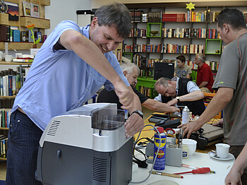 Ein Mann mittleren Alters mit blauem Hemd repariert eine Kaffemaschine. Im Hintergrund sind noch weitere Personen, die ganz unterschliedliche Geräte wie zum Beispiel einen Computer wieder auf Vordermann bringen. 