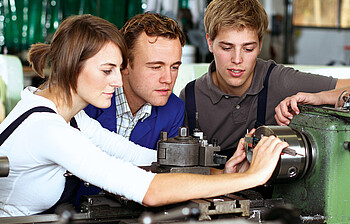 Drei junge, erwachsene Menschen, eine Frau und zwei Männer, blicken gespannt und konzentriert auf eine technische Apparatur. Sie tragen ein weißes, blaues bzw. ein braunes Oberteil. 