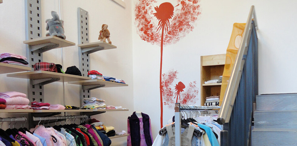 carla in Dornbirn. Ein Ausschnitt des Shops. Es hängen etliche Kleidungsstücke an Bügeln, wie es eben für ein Textilgeschäft üblich ist. Die weiße Wand im Hintergrund ziert eine rote Pusteblume. 
