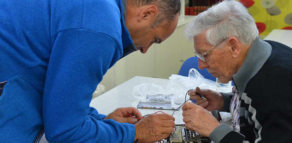 Ein älterer Herr mit grauen Haaren, einer Brille und einem schwarzen Pullover repariert mit einem Mann mittleren Alters, der ein blaues Oberteil trägt, ein Elektrogerät, indem sie Drähte anlöten. 
