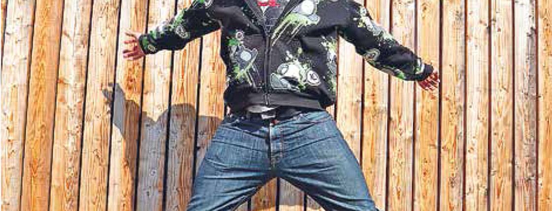 Ein junger Mann springt in die Höhe hinter einem Holzhintergrund. Er trägt eine Jeans und ein buntes Oberteil.