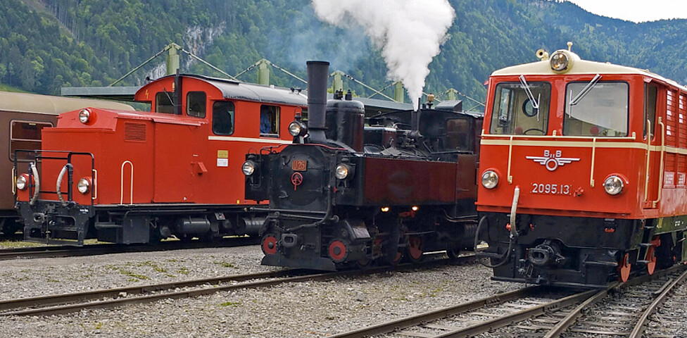 Ein gewöhnlicher Bahnhof mit einer alten, schwarzen Dampflockomotive. Auf den Gleisen daneben stehen zwei rote, etwas modernere Loks. 